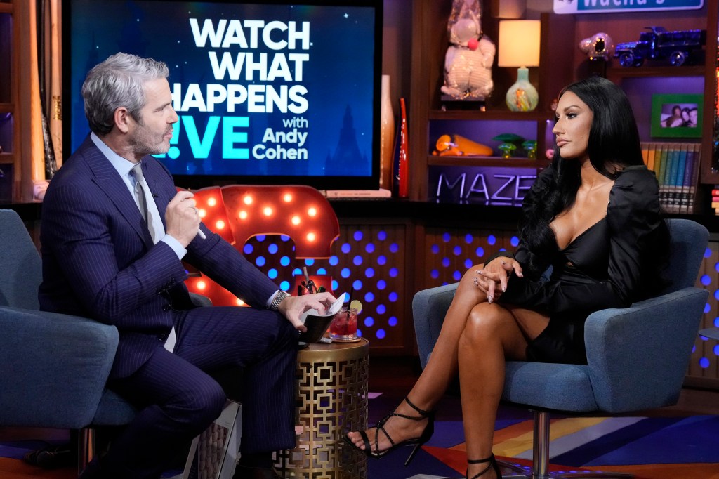 Andy Cohen in a navy blue suit on the left interviewing Monica Garcia on the right. Monica has her legs crossed and is wearing a black dress