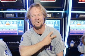 Kody Brown sitting in front of a slot machine, throwing up the peace sign and grinning