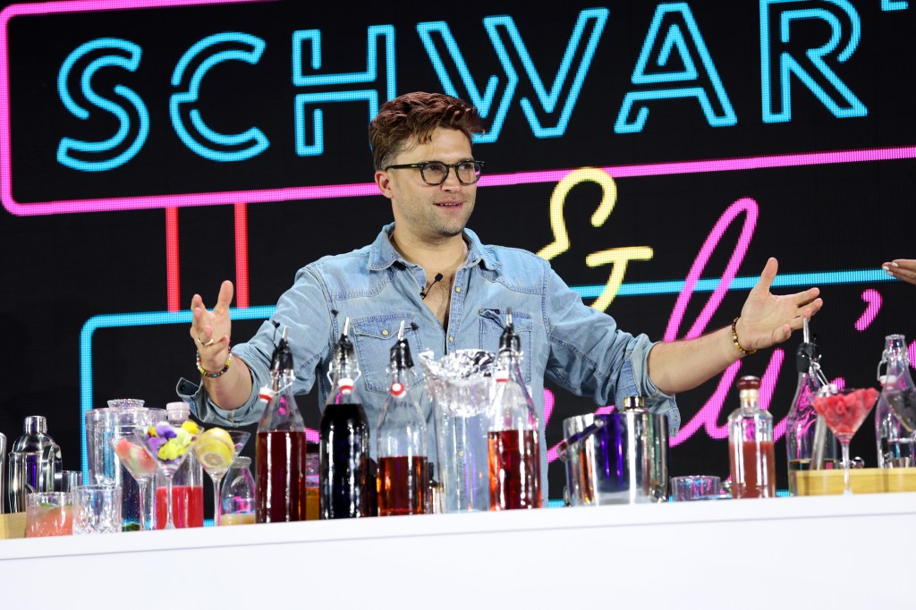 Tom Schwartz standing in front of a bar on stage at BravoCon 2022. His arms are in the air