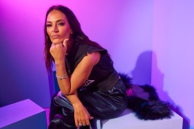 Lisa Barlow in a black outfit sitting and doing a thinking pose in front of a purple backdrop