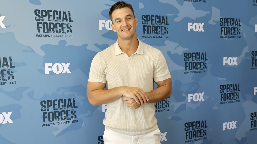 Tyler Cameron wearing white pants and a tan shirt, posing in front of a backdrop for Fox's "Special Forces."