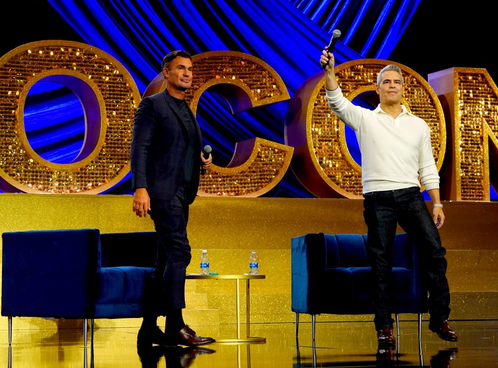 Andy Cohen in dark pants and a white sweater on stage at BravoCon 2023 holding a microphone in the air. Jeff Lewis is on the left side of the picture, wearing a suit and looking towards the audience.