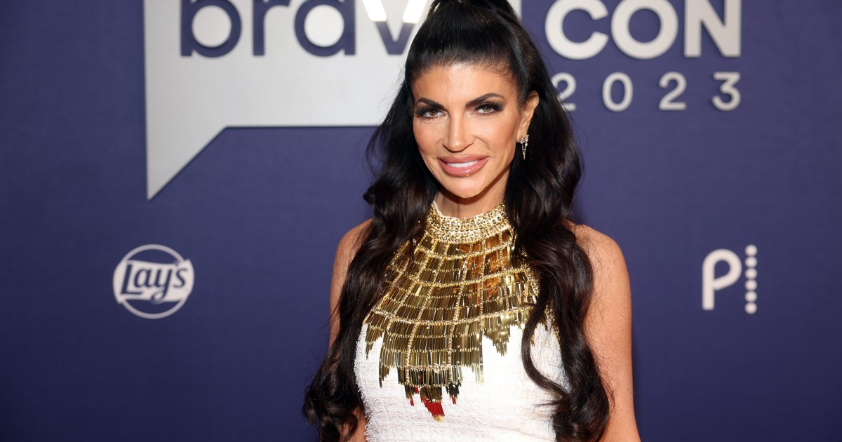 Teresa Giudice smiling and wearing a white and gold dress, standing in front of a BravoCon 2023 sign.