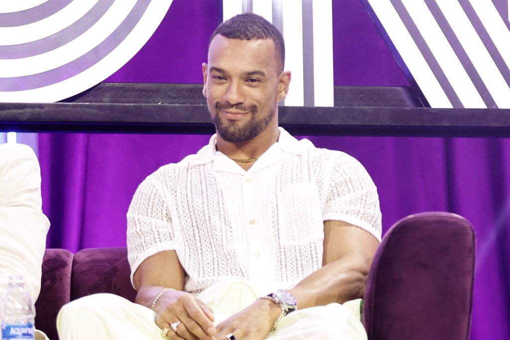 Jason Cameron in an all-white outfit sitting on stage and smirking at BravoCon 2023 surrounded by a purple backdrop