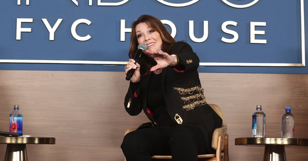 Lisa Vanderpump sitting on stage in a chair, holding a microphone and extending her hand out. She's wearing a black suit.