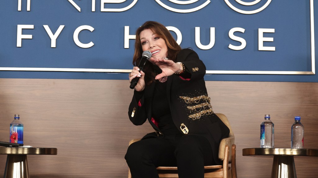 Lisa Vanderpump sitting on stage in a chair, holding a microphone and extending her hand out. She's wearing a black suit.