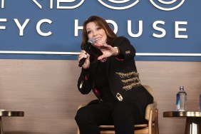 Lisa Vanderpump sitting on stage in a chair, holding a microphone and extending her hand out. She's wearing a black suit.
