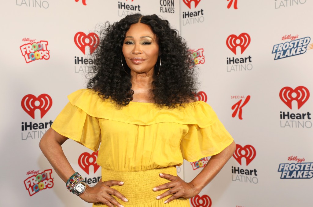 Cynthia Bailey smiling and standing with her hands on her hips, wearing a yellow dress.