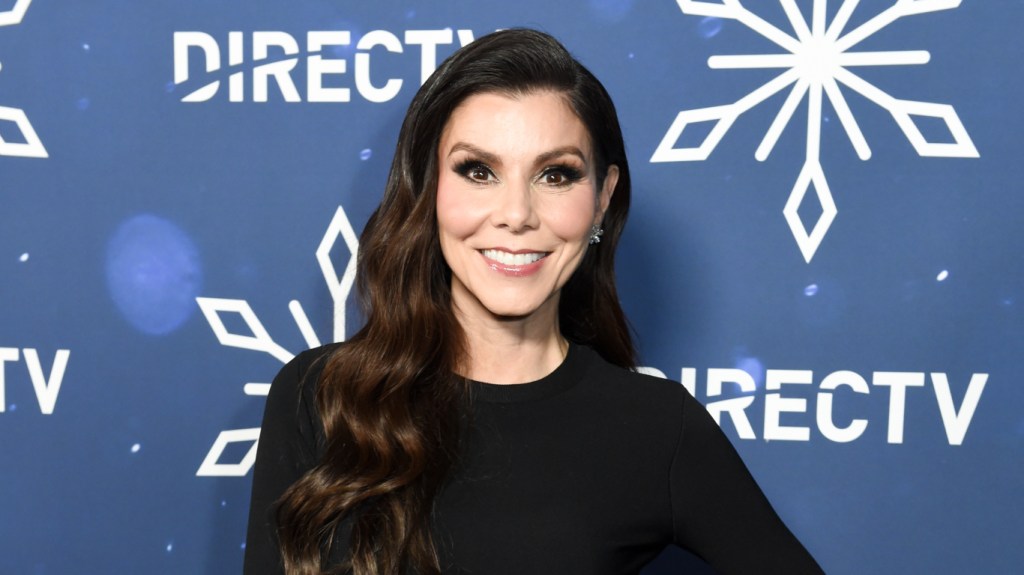 Heather Dubrow smiling and wearing a black dress, standing in front of a blue backdrop with snowflakes