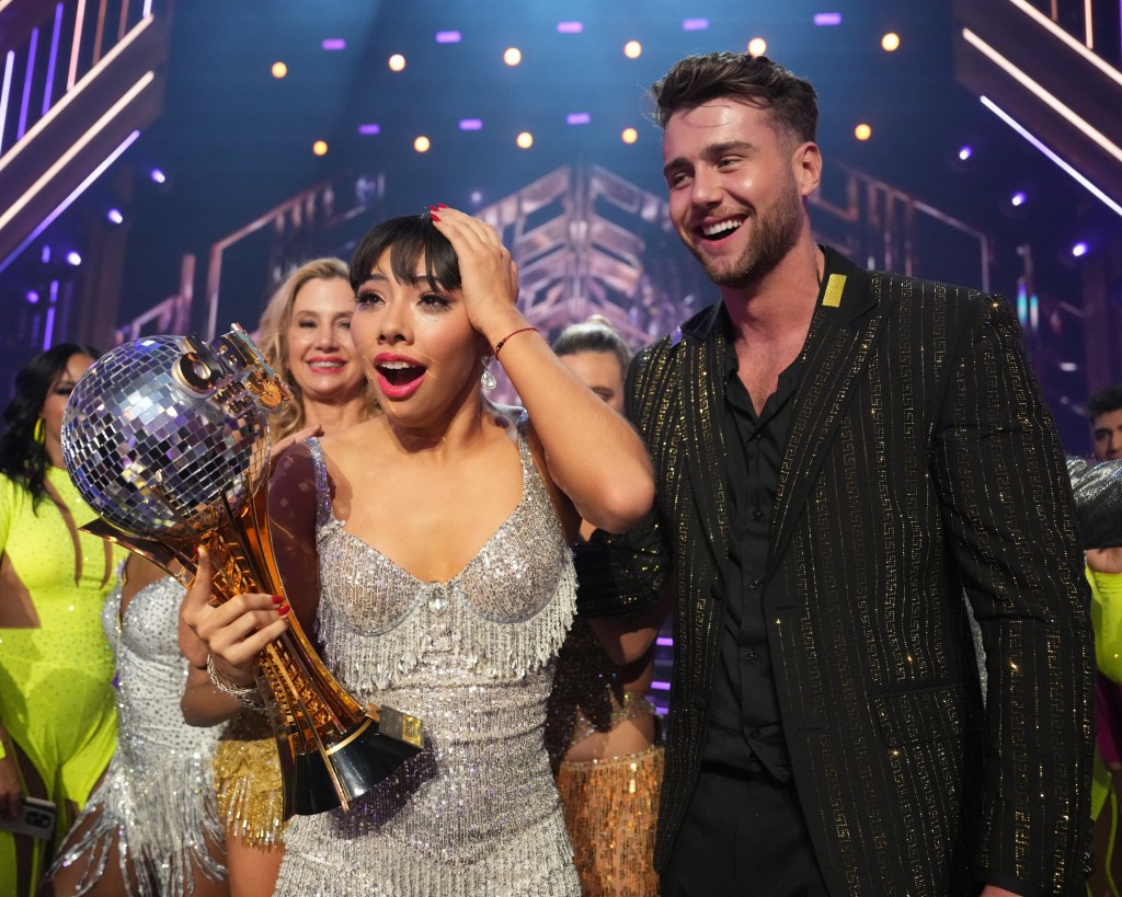 Xochitl Gomez on Dancing with the Stars reacting to being announed the winner, she's wearing a silver dress and holding the Mirrorball Trophy, standing next to Harry Jowsey who's wearing a black suit.