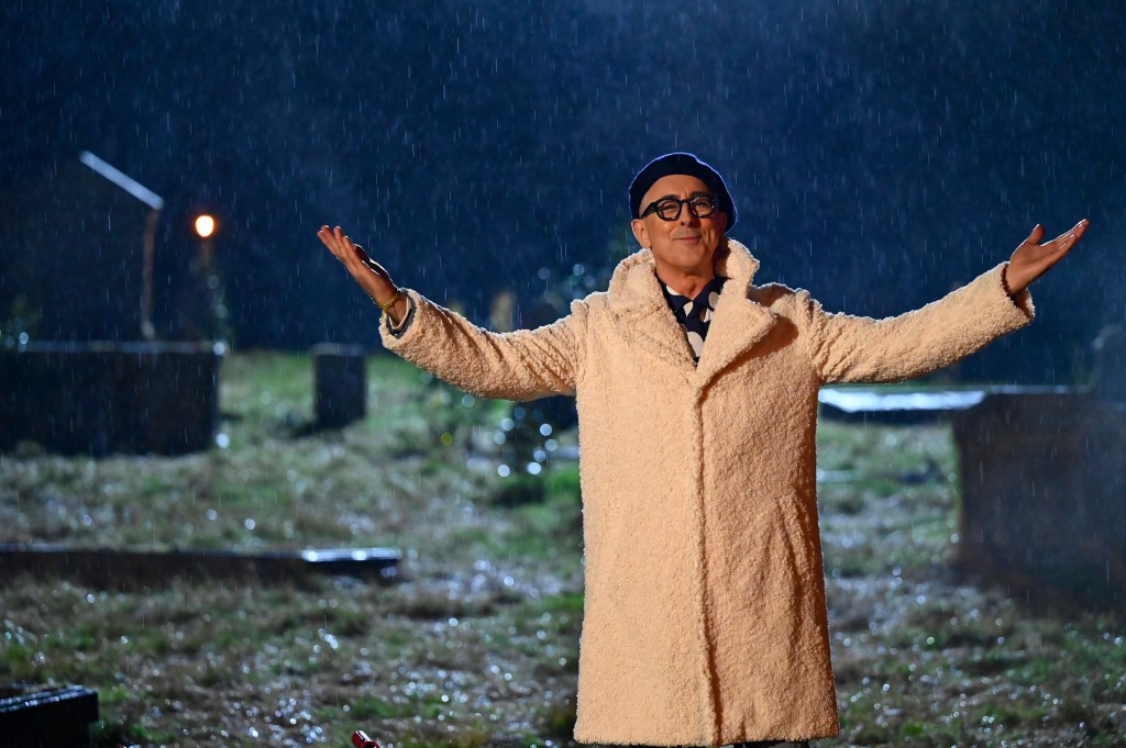 Alan Cumming in Season 2 of The Traitors, standing in a cemetery with his arms in the air and wearing a long white coat