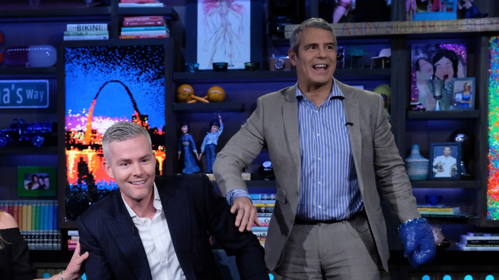 Andy Cohen standing next to Ryan Serhant who is sitting in a chair on Watch What Happens Live