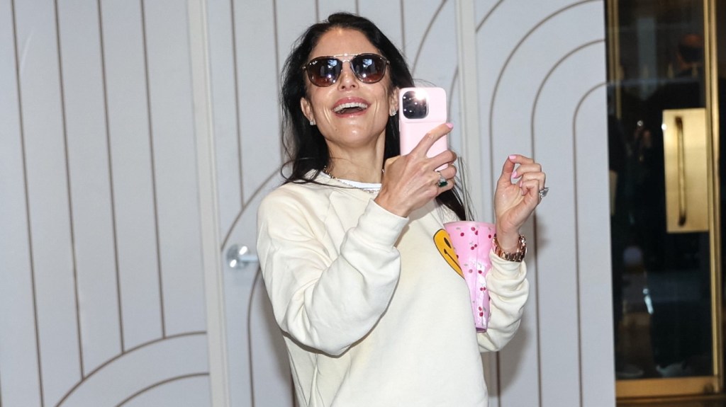 Bethenny Frankel in a white sweater and sunglasses, holding up her phone and smiling