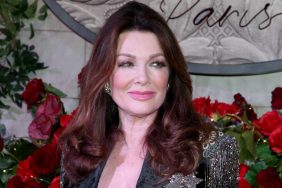 Lisa Vanderpump smiling in a black and silver blazer, standing in front of red flowers