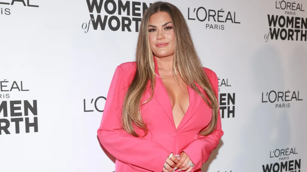 Brittany Cartwright standing and smiling in a pink blazer with her hands folded in front of her