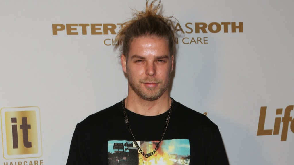 Jeremy Madix posing in front of a white backdrop wearing a messy bun and a black t-shirt