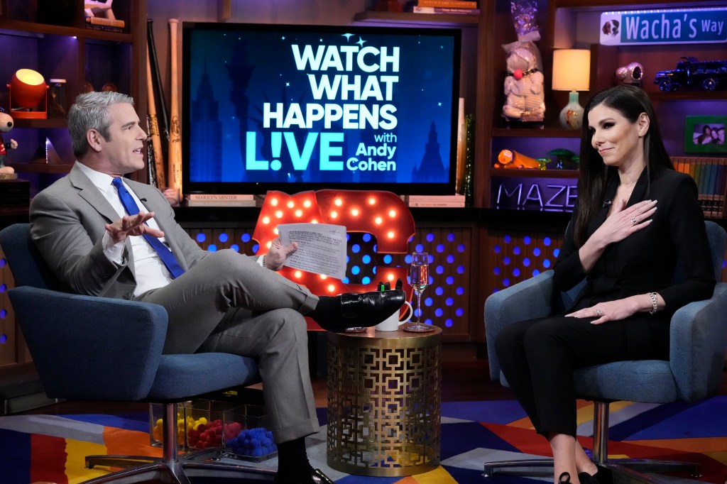 A screenshot from Watch What Happens Live where Andy Cohen is on the left in a grey suit sitting with his legs crossed, Heather Dubrow is sitting across from him in a black dress with her hand over her heart