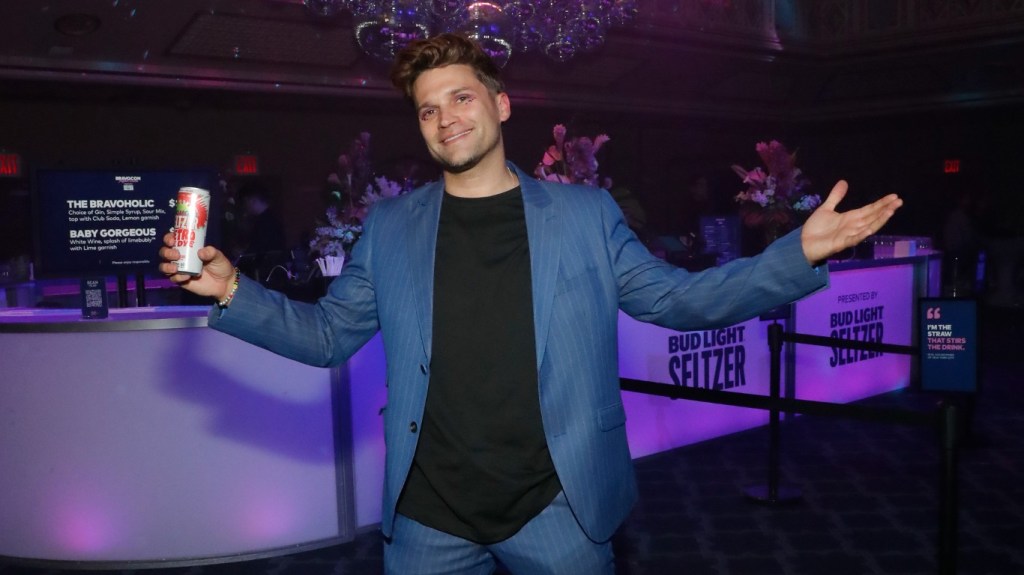 Tom Schwartz in a blue suit holding his arms up in the air while holding a canned beverage