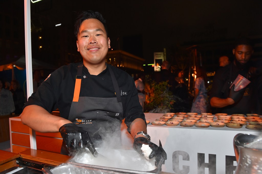 Buddha Lo in a black chef outfit on Top Chef Season 20, standing in front of a cooking range with smoke at the bottom of the image