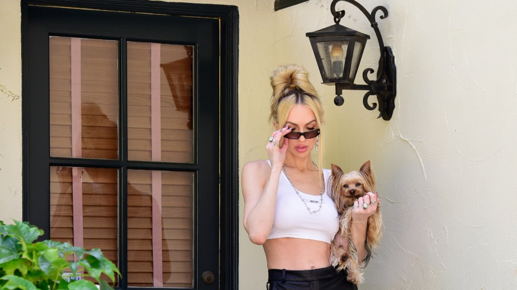 Christine Quinn standing outside of her home, covering her face with sunglasses, and holding a dog; she's wearing a white crop top