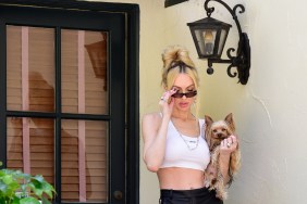 Christine Quinn standing outside of her home, covering her face with sunglasses, and holding a dog; she's wearing a white crop top