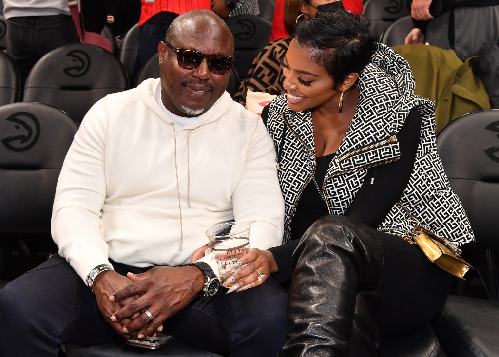 Simon Guobadia in a white sweater sitting with Porsha Williams at a basketball game, she's wearing a black and white jacket