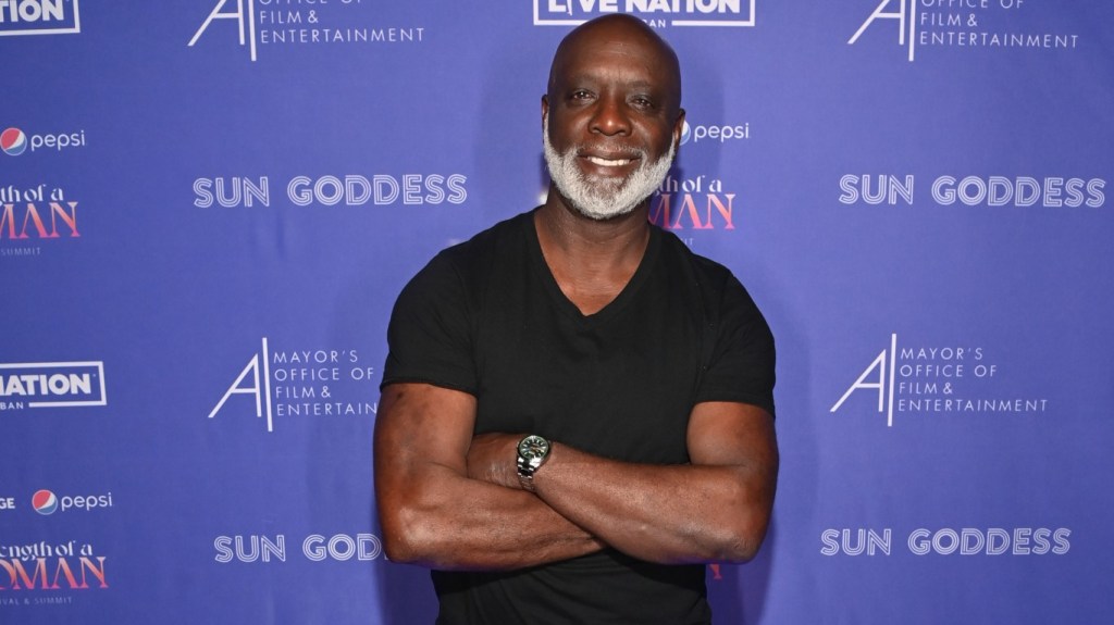 Peter Thomas in a black shirt standing in front of a blue backdrop with his arms crossed