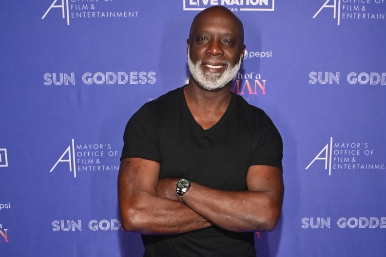 Peter Thomas in a black shirt standing in front of a blue backdrop with his arms crossed