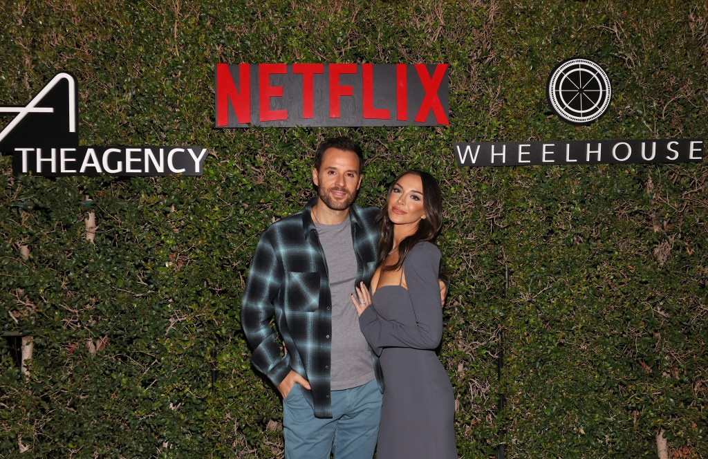 Alex Manos in a blue flannel shirt standing with Farrah Brittany, who is wearing a grey dress; they're posed in front of a bush with the Netflix and The Agency logos