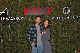 Alex Manos in a blue flannel shirt standing with Farrah Brittany, who is wearing a grey dress; they're posed in front of a bush with the Netflix and The Agency logos