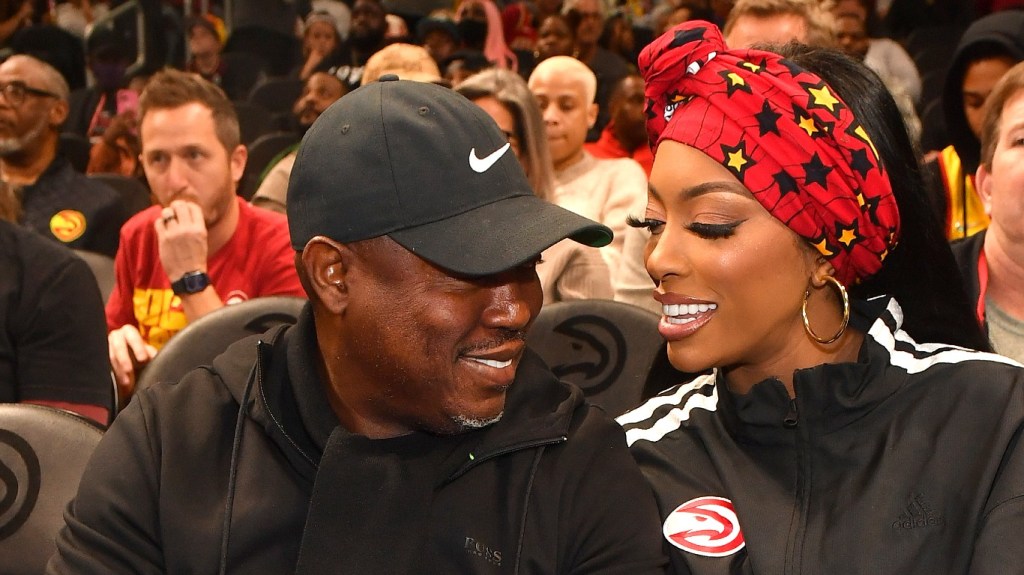 Simon Guobadia in a black jacket and black hat sitting at a basketball game and looking at Porsha Williams, who is also wearing black and a red headband