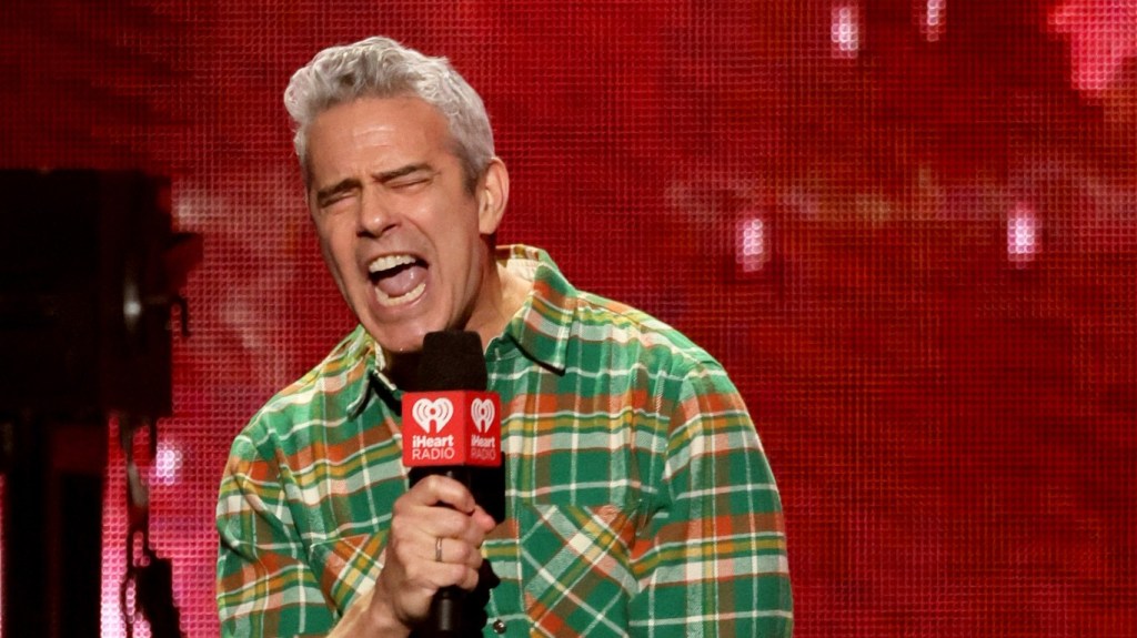 Andy Cohen in a green flannel shirt on stage screaming into a microphone with his eyes shut