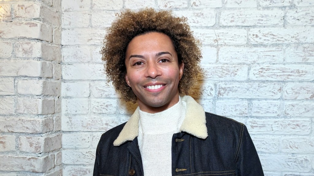 Shangela (out of drag) smiling and standing in front of a white brick wall and wearing a white sweater with a denim jacket.