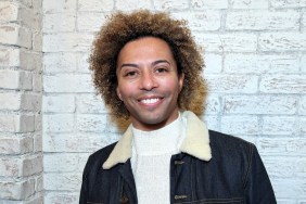 Shangela (out of drag) smiling and standing in front of a white brick wall and wearing a white sweater with a denim jacket.