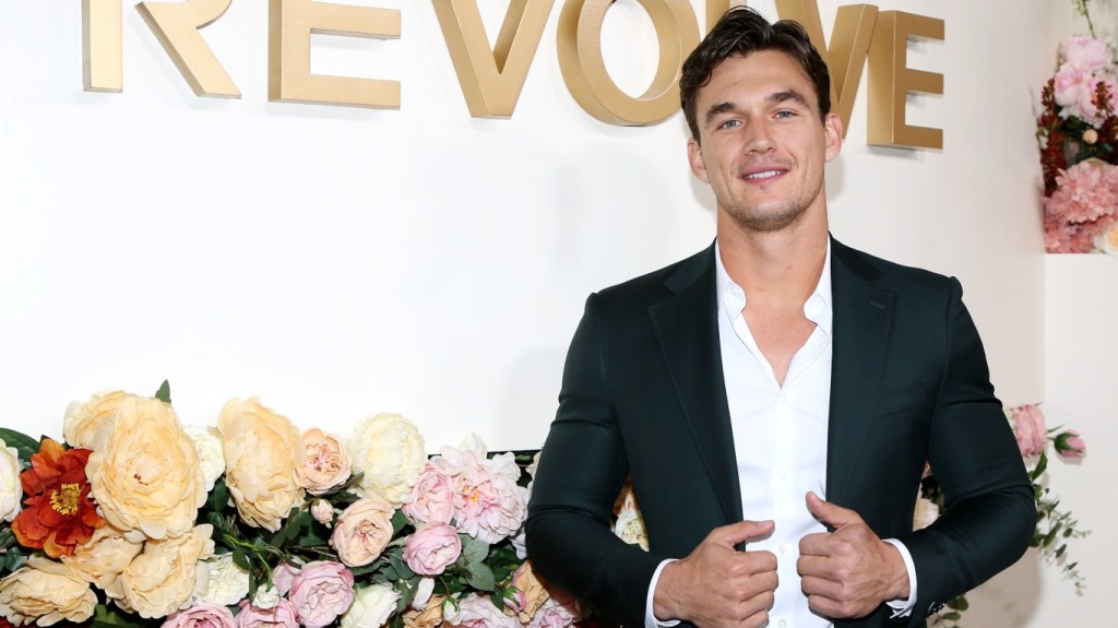 Tyler Cameron posing next to a flower arrangement, his hands are on the jacket of his open suit and he isn't wearing a tie