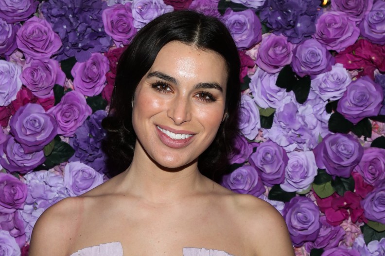 Ashley Iaconetti posing and smiling in front of a wall of purple and pink flowers