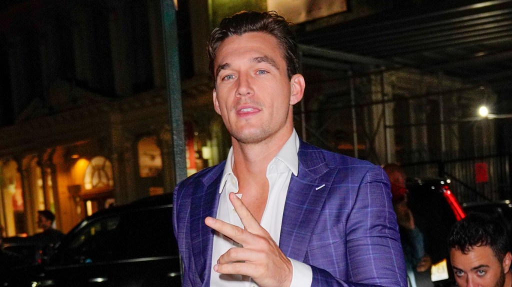 Tyler Cameron throwing up the "peace" sign and wearing an unbuttoned purple suit with his chest exposed