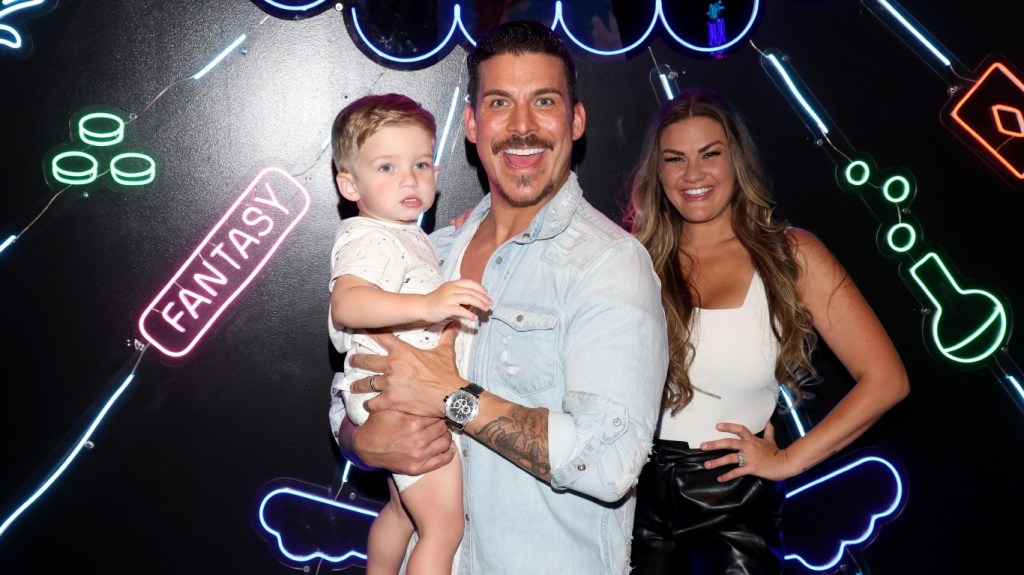 Jax Taylor and Brittany Cartwright posing together with their son, Cruz. Jax is holding Cruz and smiling