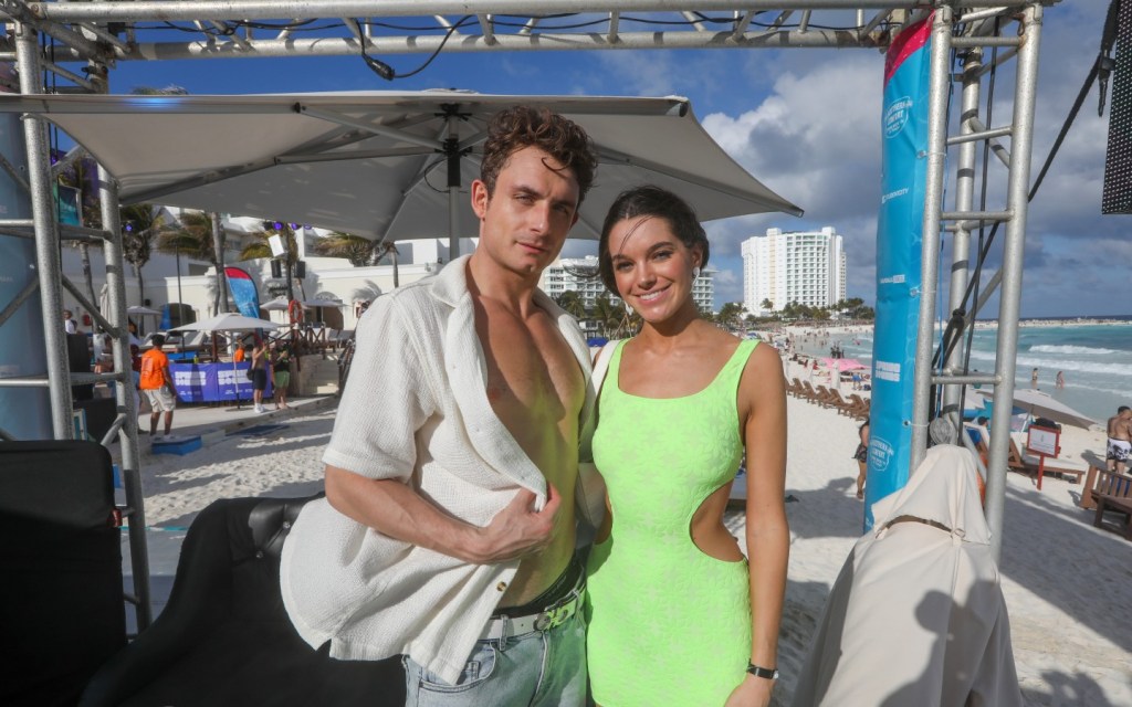 James Kennedy in an unbuttoned white shirt posing on the beach with Ally Lewber, who is wearing a green dress