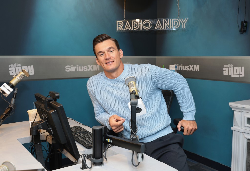 Tyler Cameron sitting in a blue sweater in a Sirius XM studio, leaning into the desk