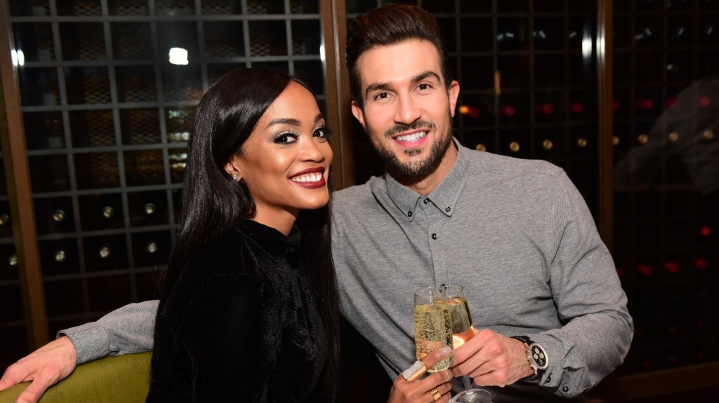 Rachel Lindsay in a black dress smiling and cheers a glass of champagne with Bryan Abasolo, who is wearing a grey shirt