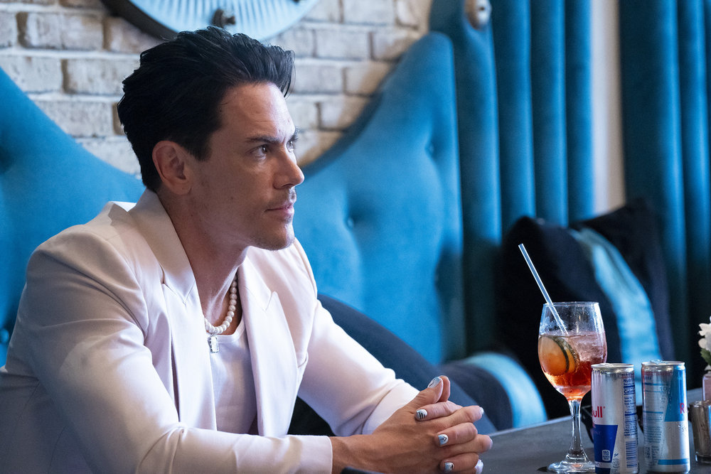 Tom Sandoval sitting in a white blazer at a table with drinks scattered across it