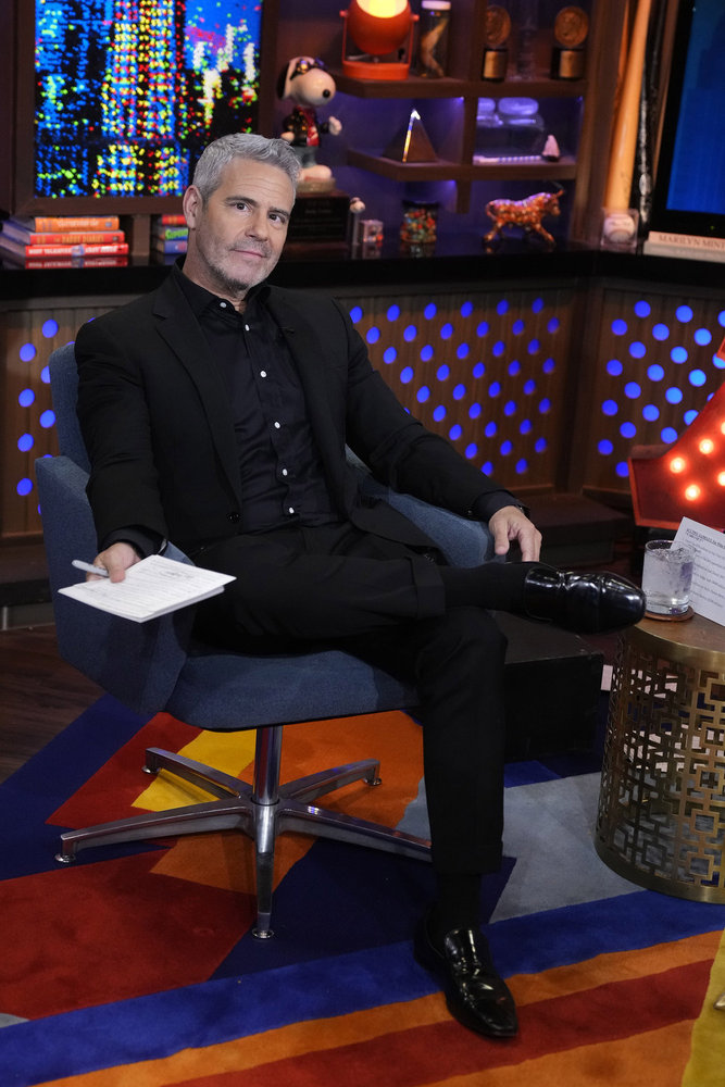 Andy Cohen in a black suit sitting with his legs crossed on Watch What Happens Live
