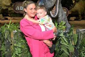 Brittany Cartwright in a pink shirt holding Cruz and standing in front of an animatronic dinosaur