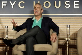 Andy Cohen sitting with his legs crossed in a beige chair while wearing a blue shirt and black suit