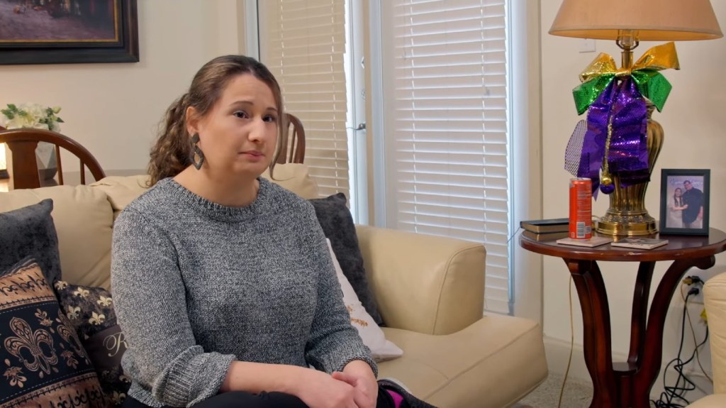 Gypsy Rose Blanchard sitting on a couch in a grey sweater making a contemplative expression