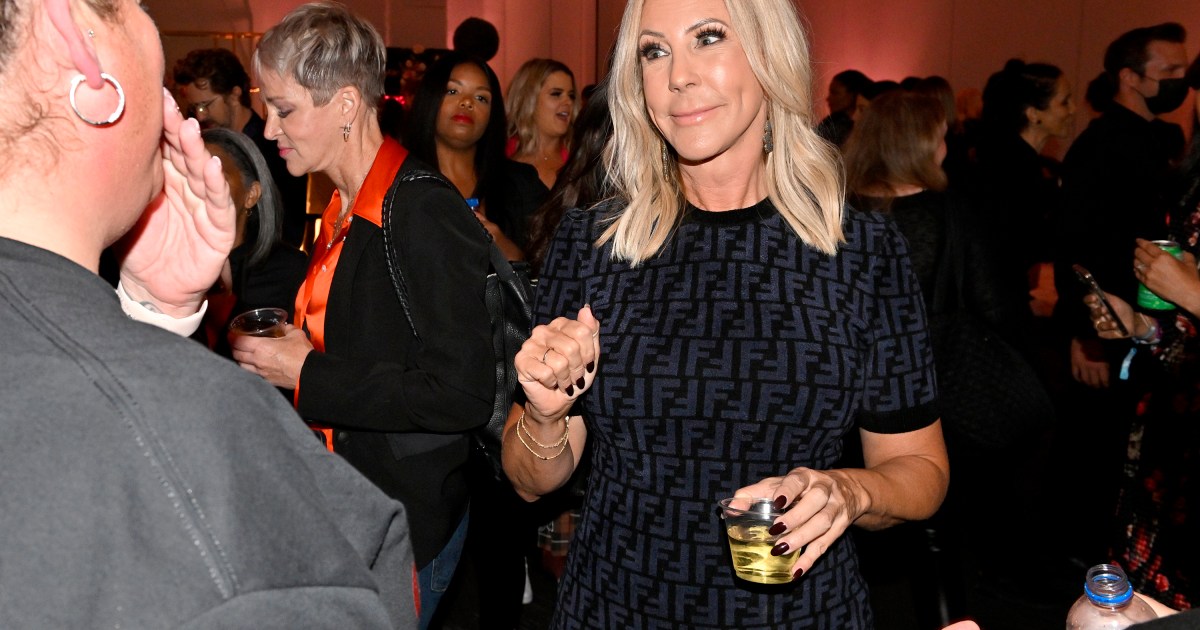 Vicki Gunvalson in a sea of people, smiling, wearing a black dress, and holding a cocktail