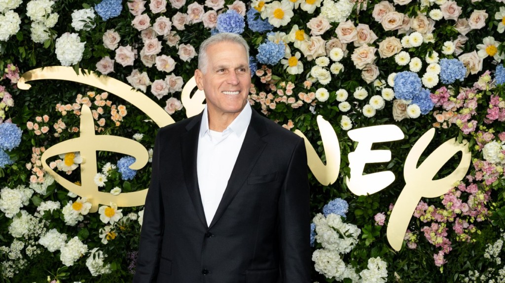 Mel Owens in a black suit posing in front of the Disney logo and a wall of flowers