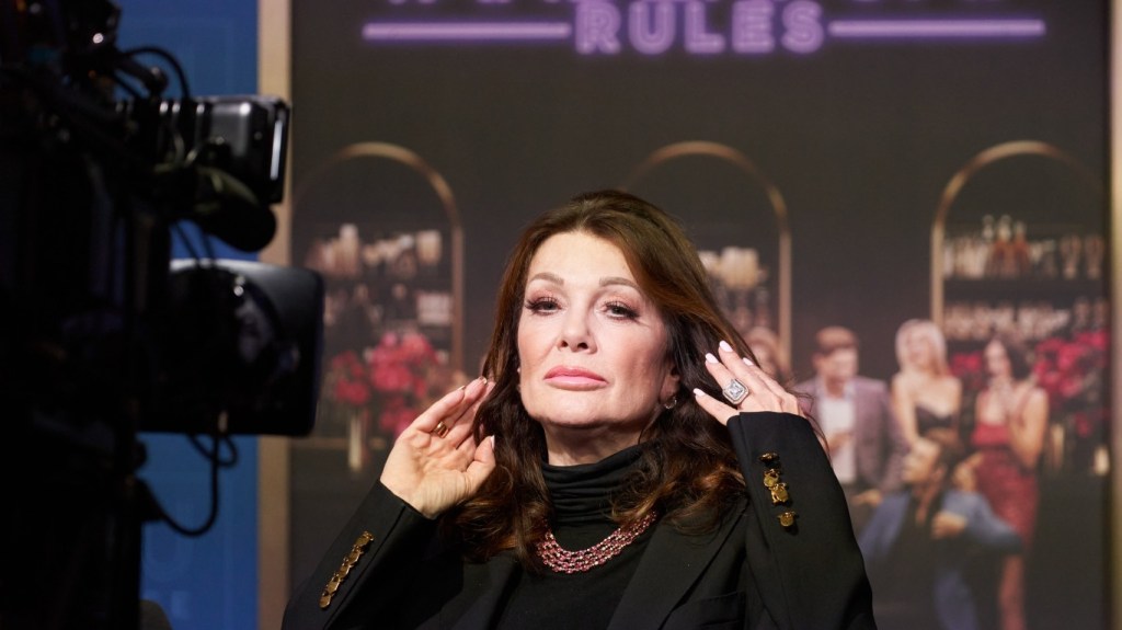 Lisa Vanderpump posing in front of a camera and a Vanderpump Rules poster