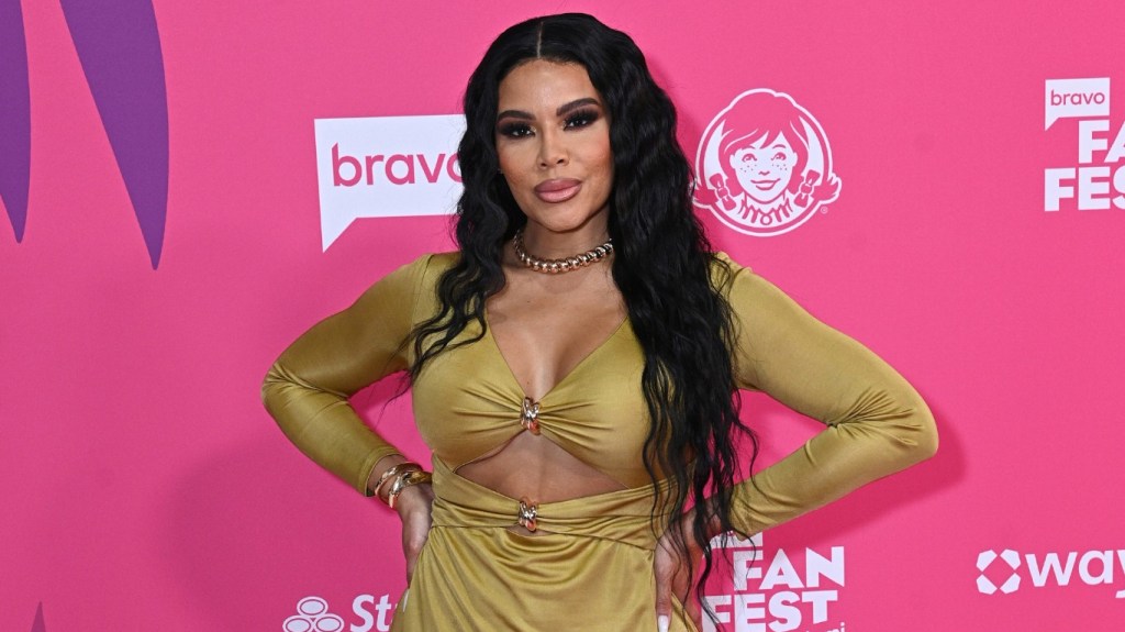Mia Thornton in a yellow dress posing with her hands on her hips in front of a pink backdrop